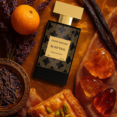 Perfume bottle labeled 'Sticky Berries by Khyal' surrounded by vanilla beans, orange, lavender, and a pastry on a brown surface.
